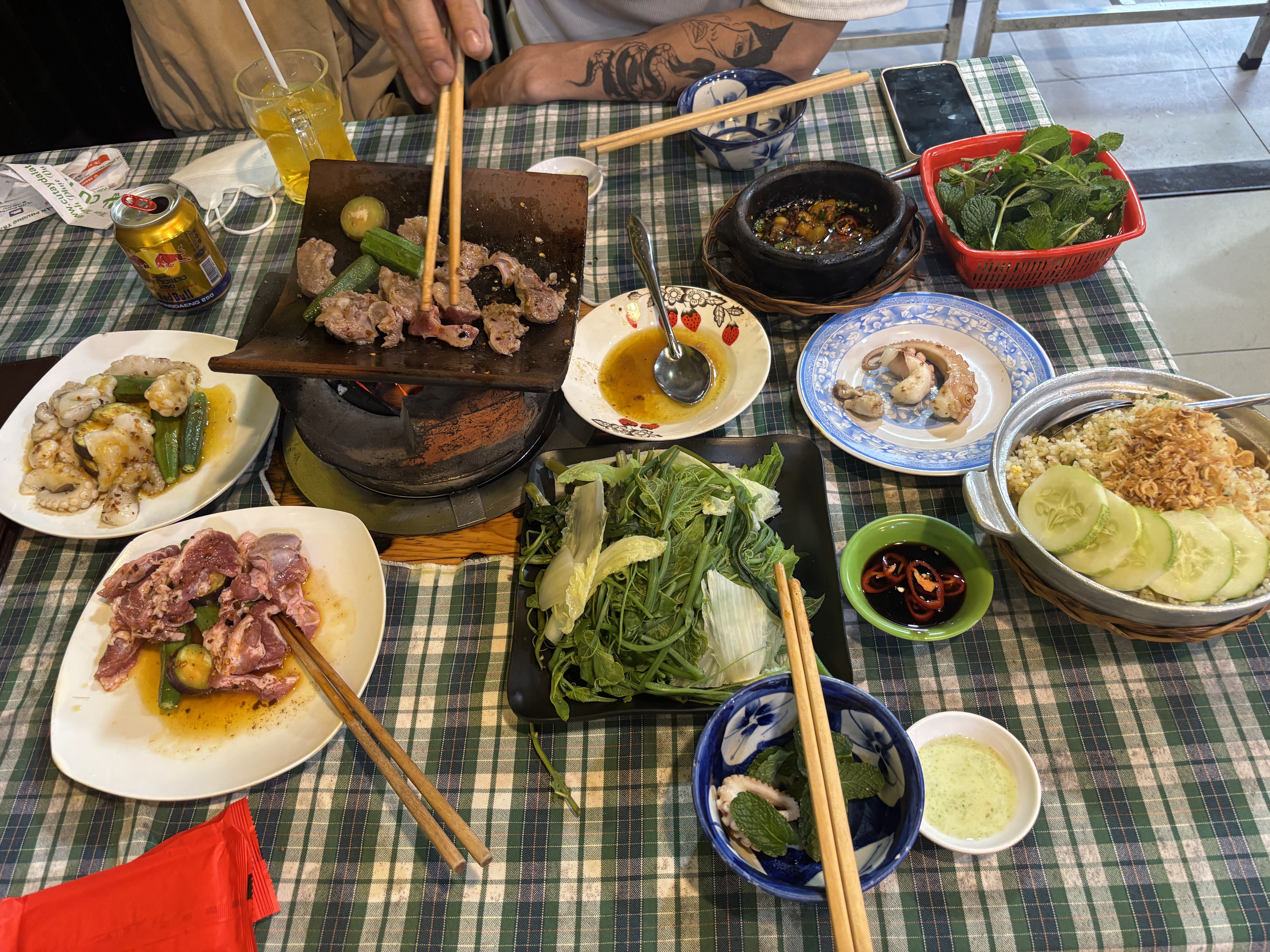 Bàn ăn phủ khăn caro với bếp nướng ngói, thịt, hải sản, rau xanh, cơm chiên và nước chấm